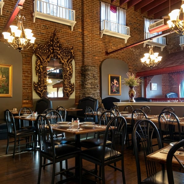 Restaurant dining room with brick walls, ornate mirrors, chandeliers, and set tables with black chairs.